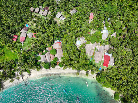 AERIAL TOP DOWN: Seaside village on tropical Masokut Island hidden in a dense palm forest. Wooden longhouses and huts line a sandy shore with clear turquoise water and small boats floating near beach.