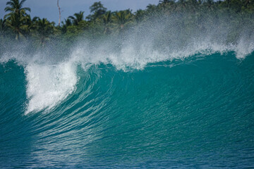 Naklejka premium CLOSE UP: Blue wave rises and folds with heavy white spray near a tropical island shoreline. Texture, shape, and force of the ocean against an Indonesian coastline overgrown with lush palm trees.