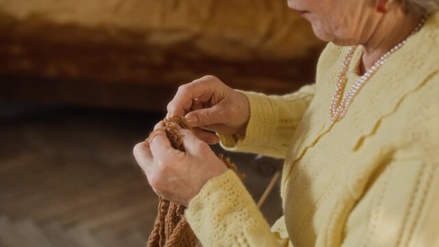 Close up on hands of unrecognizable elderly woman knitting wool scarf with precision using needles while working on handcrafted garment in bedroom