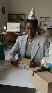 Vertical view of African American gen Z girl wearing party hat opening gift box gaping in astonishment while celebrating her birthday at workplace