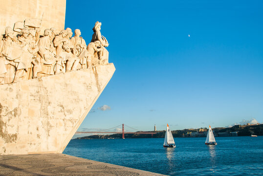 Visitors admire the Monument to the Discoveries on the Tagus River in Belem, Lisbon, while sailboats navigate the waters nearby