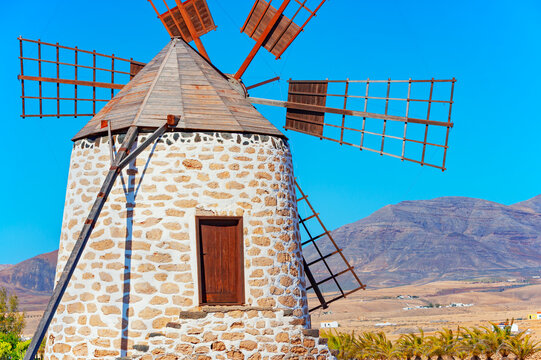 Traditional windmill stands in Fuerteventura under bright sun with clear blue sky and mountains in background