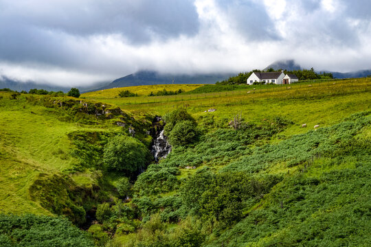 Cottage and waterfall stand in green landscape near Staffin on the Isle of Skye in Scotland surrounded by hills and clouds