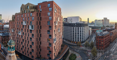 A sweeping aerial panorama of Whitworth St in Manchester as the golden hour sun illuminates modern brick high-rises and the city's urban landscape. © bardhok