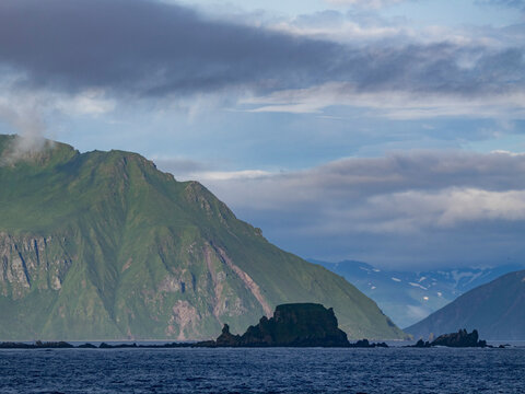 Views of the rocky Baby Islands in the Aleutian Islands of Alaska with mountains in the background and cloudy skies above