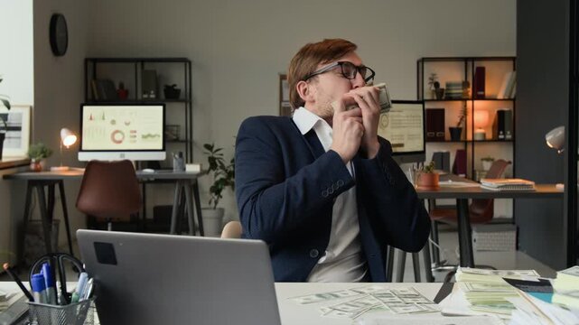 Zoom in shot of overjoyed gen Z man with glasses in formalwear holding stack of dollar bills and kissing it while sitting at office desk