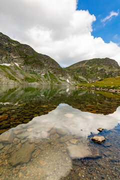 Clear mountain lake with rocks visible in Rila Mountains Bulgaria under partly cloudy sky and snow patches on slopes