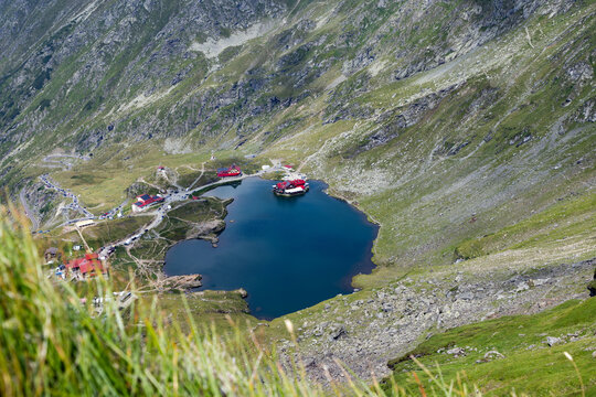 View of Balea Lake in Fagaras Mountains during a sunny day with green hills surrounding the blue water in Romania