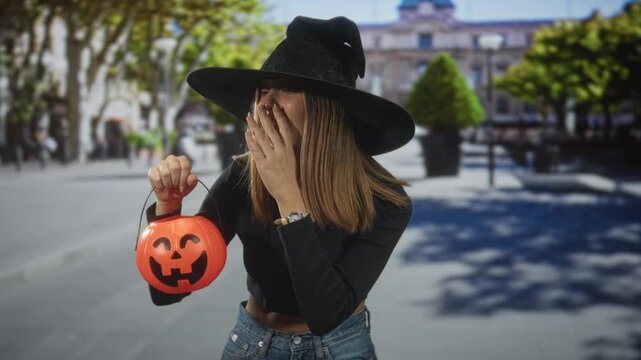 Woman in witch hat holding jack o lantern with hand to mouth gesture on a street in daylight; festive surprise.