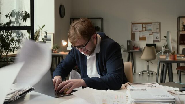 Overloaded gen Z office worker finishing project on laptop dealing with tight deadline and dodging papers flying toward him while he sitting at desk