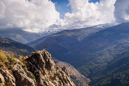 Snow-capped Sierra Nevada mountains and cross on rock viewed from mountains in Guejar Sierra, Granada province, Spain under bright clouds
