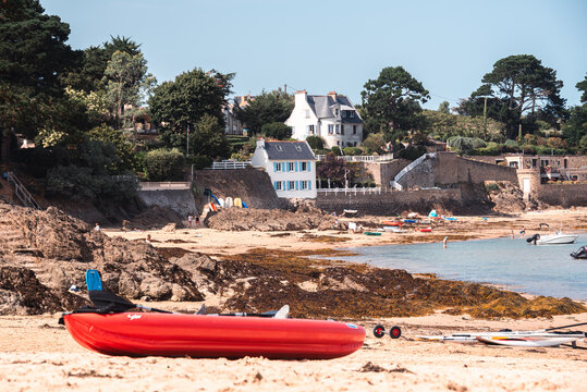 Bright red canoe on rocky beach with coastal houses and pine trees in Rotheneuf France