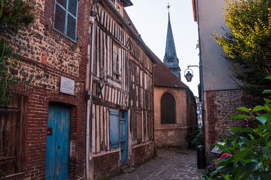 Narrow cobbled street features medieval houses and a church spire in the historic district of Honfleur in France