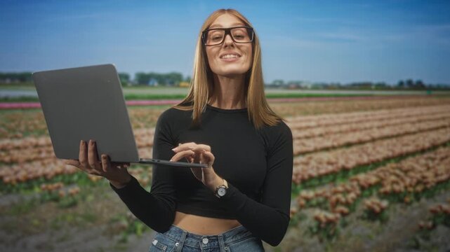 Redhead woman holding laptop and typing with finger on trackpad while smiling in a tulip field during spring sunlight; remote work confidence.