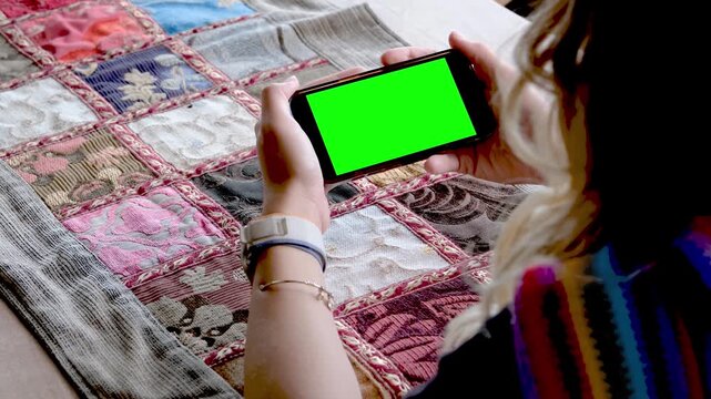 Close up of female hands holding a smartphone with green screen chroma key in landscape mode for watching movies or mobile game demonstration while sitting at a colorful table