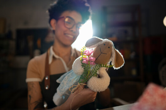 A woman smiles as she holds a doll made of fabric, adorned with flowers. She stands in a cozy room filled with various crafts and decorations, enjoying a moment of joy and creativity.