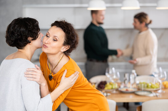 In cozy home atmosphere, two women hug and kiss on cheek, say goodbye after party. Their boyfriends are in background.