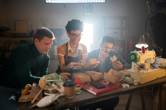 Three people gather around a table in a workshop. They look engaged as they work on small craft items. Tools and materials are scattered on the table. The lighting is warm.