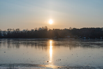 Naklejka premium A tranquil frozen lake scene, partially covered with ice and snow The low sun casts a warm glow across the barren tree expanse under a clear sky, reflecting off the ice Colors transition from vibra