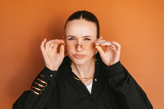 Young stylish woman squinting eyes to see better or looking with suspicion, holding yellow glasses with both hands, curious female model in black shirt against orange studio background, close up
