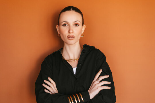 Portrait of confident young stylish woman looking at camera with crossed arms, female model with slicked back hair and gold jewelry, wearing black shirt, isolated on orange terracotta background
