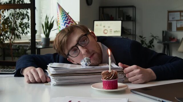 Apathetic young man wearing party hat and glasses resting head on stack of papers and blowing into party horn, with cupcake and lit candle put before him during somber birthday celebration at work
