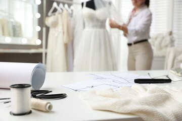 Naklejka premium Dressmaker working with wedding dress on mannequin in atelier, focus on white table with tools, fabric and patterns