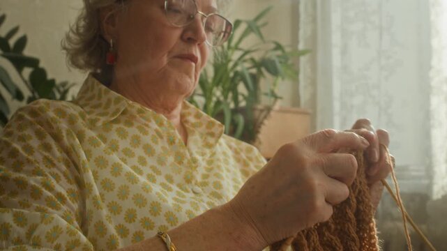 Tilt up shot of focused older woman wearing eyeglasses working on wool scarf crafting garment with needles while spending relaxing afternoon at home
