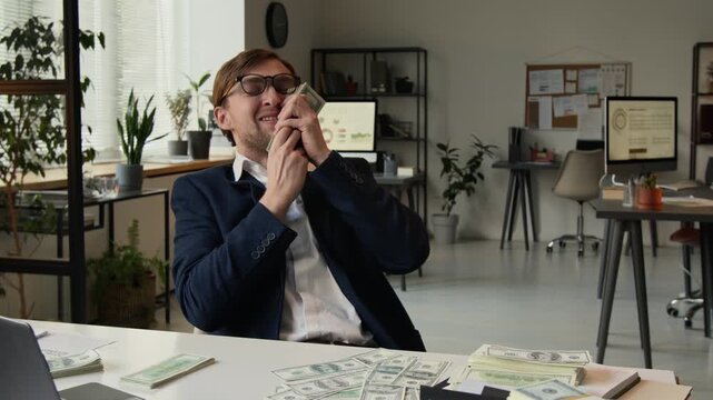 Delighted young man flipping through stack of money, kissing it with smile and adjusting glasses at workplace