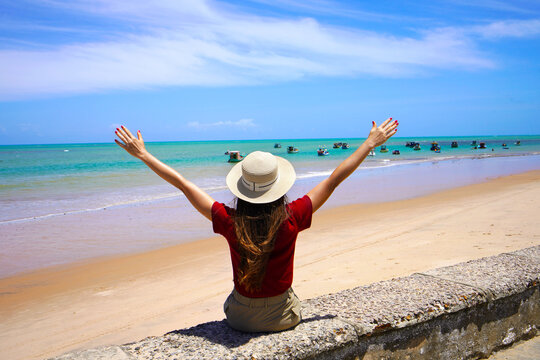 Traveler woman sitting on wall with open arms enjoying sight of natural swimming pools with jangadas rafts in Joao Pessoa, Brazil