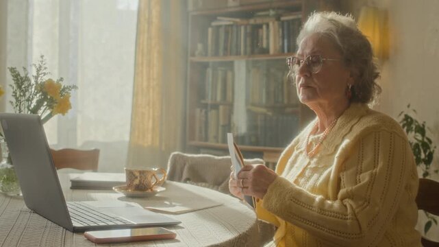Medium shot of elderly woman with grey hair typing on laptop then opening envelope with electricity bill while reviewing financial charges online at table in retro apartment