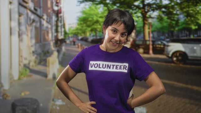 Woman in volunteer tshirt standing with hands on hips and wiping forehead on a tree lined street near parked car and sidewalk; determination.