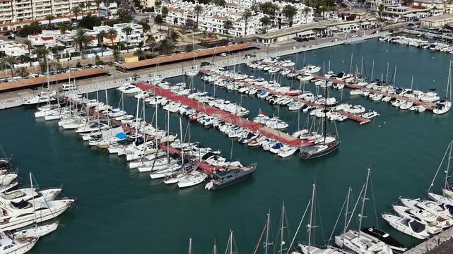 Many boats are parked at a marina with buildings and trees in the background. The scene shows the hustle of a busy coastal town during daytime.