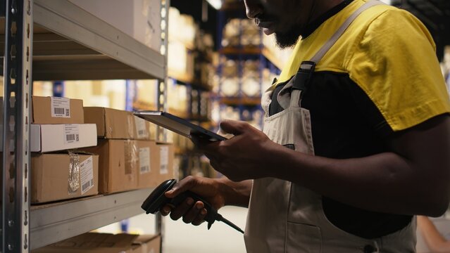 African american e-commerce employee scanning awb tracking numbers, registering shipping labels and details for the warehouse inventory records. Handling pallets import export operations. Camera A.