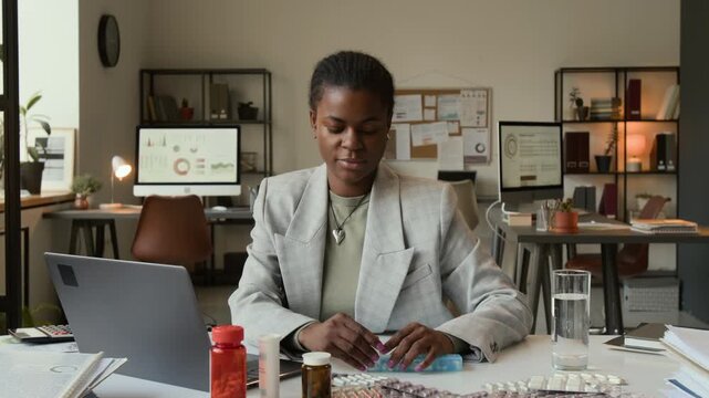 Zoom in view of African American gen Z female manager opening plastic pillbox, taking pill and drinking it with water while working on laptop at white office desk