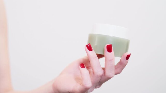 Woman holds a small jar of cream with green contents in her right hand in a bright indoor space during the day