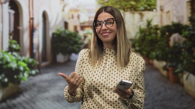 Woman wearing glasses and patterned blouse holding smartphone points thumb with left hand on cobblestone street while smiling; friendly confidence.