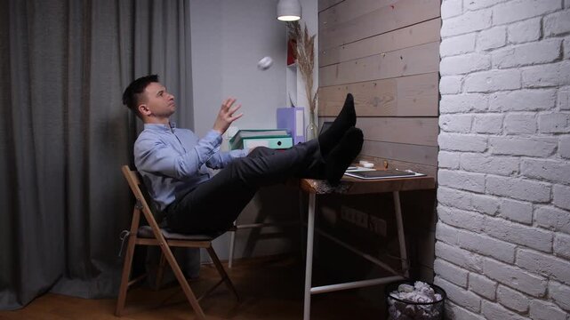Young man in a home office sits with feet on desk, procrastinating work by tossing a crumpled paper ball, embodying distraction, boredom and unproductivity during remote work