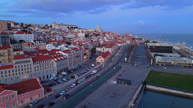Drone camera shows multilane waterfront road, parked cars, small marina docks and layered residential blocks rising on hillside beside calm river water under soft evening sky