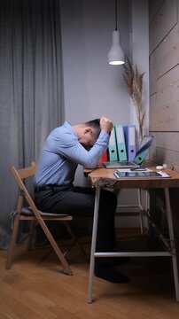 Vertical video frustrated man sitting at home office desk, holding his head in despair, feeling overworked and suffering from overwhelming stress and anxiety