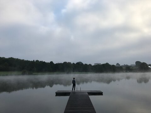 Moody and atmospheric lakeside scene featuring a solitary person standing on a wooden pier extending into a calm lake covered with soft mist. The water reflects surrounding trees that fade into fog.