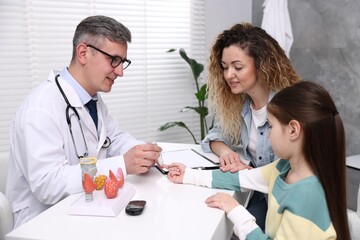 Obraz premium Endocrinologist in glasses checking little girl's blood sugar level with lancet pen while her mother supporting her at white desk in clinic