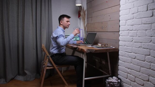 Man working remotely on a laptop at a rustic wooden desk, staying hydrated by drinking water while focusing on his professional tasks in a modern home office