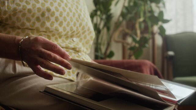 Close up on hands of elderly woman turning pages of family photo album on lap looking through retro pictures while remembering heartwarming life events in living room
