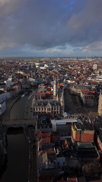 Aerial Ghent Belgium cityscape under dramatic clouds, cinematic drone sweep over medieval rooftops, canals and belfry warm sunlight slicing through cloudbreaks panoramic reveal of cobblestone