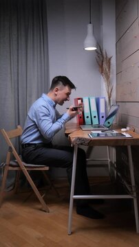 Vertical video stressed man communicating with anger and frustration on a mobile phone during a remote work call, sitting at a rustic wooden desk with a laptop and documents