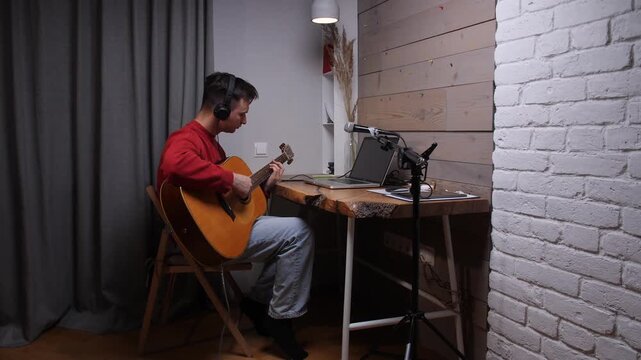 Man wearing headphones and red sweater playing acoustic guitar, sitting at a wooden desk with a laptop and microphone, recording music in a comfortable home studio setup