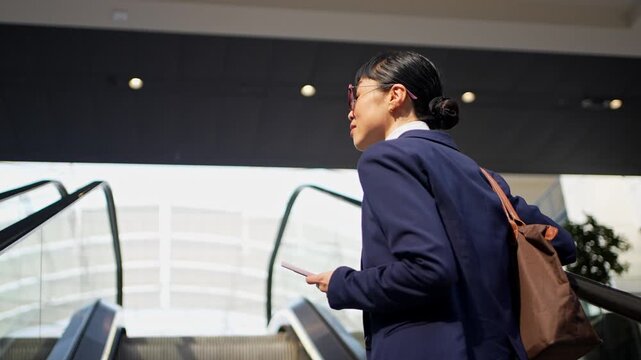 Young Japanese businesswoman riding escalator with phone and bag