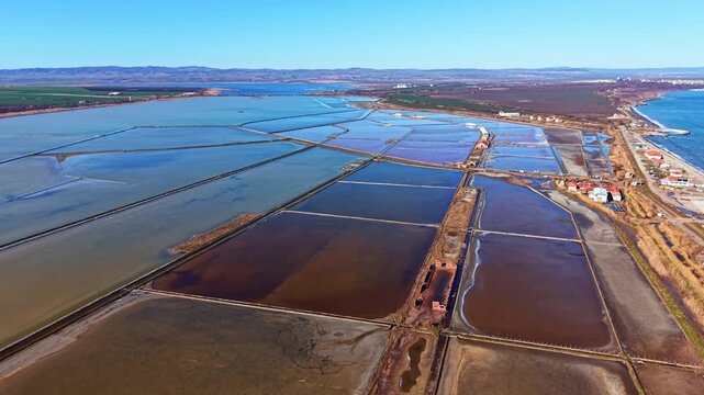 Large salt flats cover the land near the shore. Water pools reflect the bright blue sky. The area appears divided into sections. The coastline is seen in the distance.
