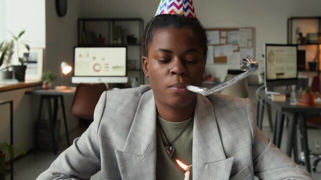 Tilt up view of apathetic Black young woman wearing birthday hat blowing into party whistle when sitting in front of small plate with cupcake and burning candle in modern office